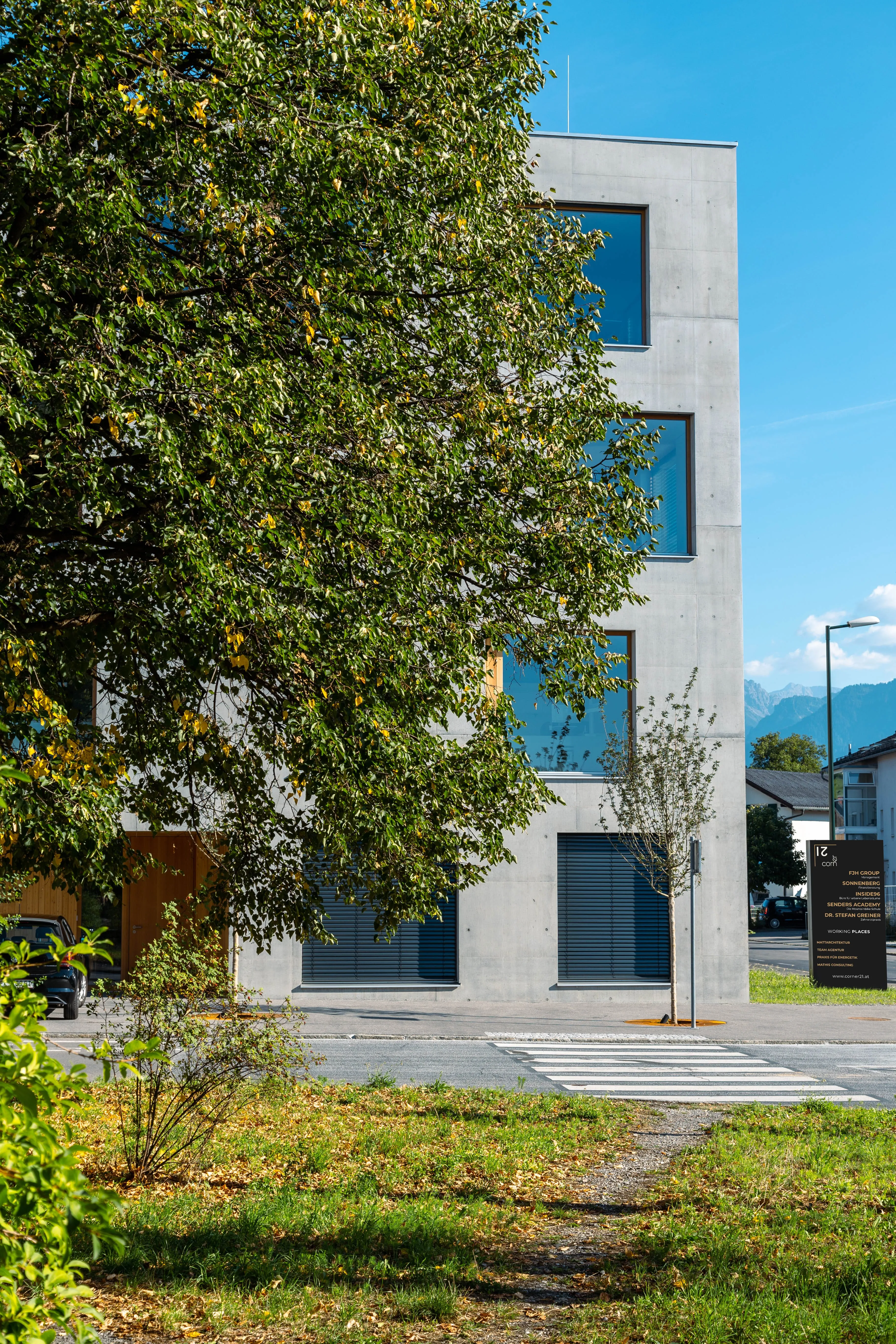 Corner 21 in Rankweil: Sichtbetongebäude hinter Bäumen mit Fußgängerübergang.